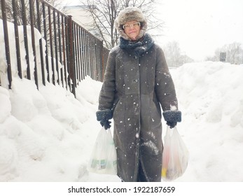 Older Mature Woman Carries Shopping Bags In A Heavy Snowfall. Middle Aged Grandmother walking Out Of The Store In A Blizzard. The Concept Of Bad Weather In The City