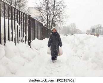 Older Mature Woman Carries Shopping Bags In A Heavy Snowfall. Middle Aged Grandmother walking Out Of The Store In A Blizzard. The Concept Of Bad Weather In The City