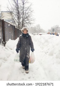 Older Mature Woman Carries Shopping Bags In A Heavy Snowfall. Middle Aged Grandmother walking Out Of The Store In A Blizzard. The Concept Of Bad Weather In The City