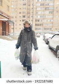 Older Mature Woman Carries Shopping Bags In A Heavy Snowfall. Middle Aged Grandmother walking Out Of The Store In A Blizzard. The Concept Of Bad Weather In The City