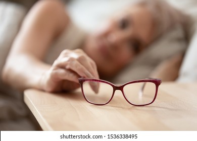 Older mature woman holding taking optical glasses from bedside table waking up in morning, old people eye sight problem, optometry optics ophthalmology, bad vision concept, eyeglasses close up view