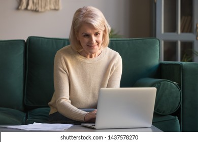 Older Mature Woman Using Wireless Laptop Apps Browsing Internet Sit On Sofa, Smiling Middle Aged Grandmother Working Distantly On Computer Surfing Web Communicating Online Looking At Screen At Home