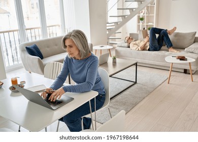 Older Mature Woman Wife Remote Working Online From Home Office, Watching Webinar Using Laptop Computer While Her Senior Old Husband Relaxing On Couch In Living Room. Middle Aged Family Couple At Home.