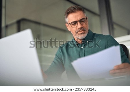Older middle aged busy professional business man executive manager or entrepreneur working on laptop computer in office checking legal corporate paperwork, reviewing financial document management.