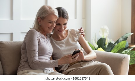 Older mother and adult daughter hugging, using phone together, young woman and mature mum, grandmother looking at smartphone screen, watching video or photos, sitting on cozy sofa at home together