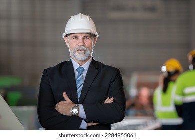 older owner engineer with beard mustache on face standing smile with his arms crossed at construction factory site. Boss manager in suit show good thumb at factory industry site. - Powered by Shutterstock