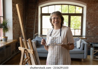 Older Painter Woman Posing Near Easel With Canvas Holds Palette And Paintbrush Smile Look At Camera Feel Inspired, Enjoy Favourite Creative Hobby In Loft Workshop. Concept Of Professional Occupation