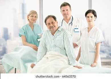 Older Patient Sitting On Bed With Hospital Crew In Background.