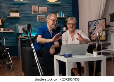 Older People Waving At Video Call Camera On Laptop While Sitting Together On Living Room Sofa. Senior Couple Talking On Online Remote Conference Having Crutches And Walk Frame On Couch