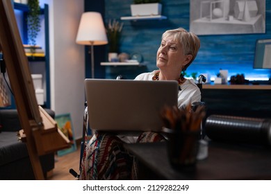 Older Person Sitting In Wheelchair Holding Laptop Looking At Pencil Sketch Drawing On Easel. Elderly Woman Artist Living With Disability Using Portable Computer Looking For Inspiration.