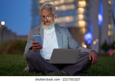 Older senior adult business man professional, mature old businessman sitting outside office holding cell phone using smartphone and laptop working online on technology devices late in night city park. - Powered by Shutterstock