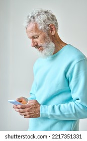 Older Senior Bearded Stylish Hipster Man Holding Cell Phone Looking At Cellphone Using Smartphone Mobile Application Buying Ordering Goods Online Standing Isolated On White Wall. Vertical