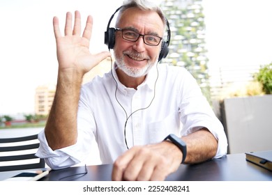 Older senior business man, online teacher wearing headphones looking at webcam waving hand teaching or elearning virtual class, having hybrid remote video call meeting concept. Web camera view shot - Powered by Shutterstock