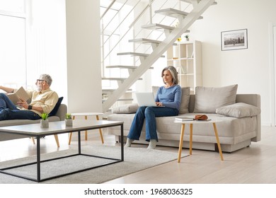 Older Senior Family Couple Spending Time At Home In Modern Living Room. Middle Aged Husband Reading Book Relaxing Sitting On Couch While Mature Wife Using Laptop Enjoying Casual Daily Activities.