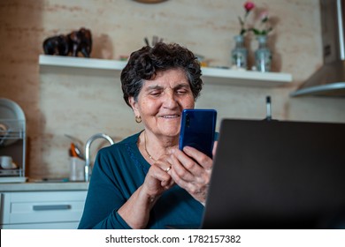 An Older Senior Woman With Black Short Hair Is Sitting In The Dining Room Laughing And Looking At The Smartphone That Is In Her Hand. It Is Located In Front Of The Laptop.