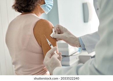 Older Senior Woman In Facemask Getting Covid19 Protective Vaccine Receiving Antiviral Injection In Arm In Clinic Hospital With Doctor. Vaccination Coronavirus Safety Concept. Back View.