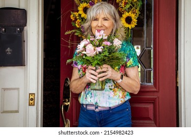 Older silver haired woman in jeans and floral tee-shirt  smiling at residence entrance door with leaded window and sunflower wreath holding just delivered pink flowers in vase