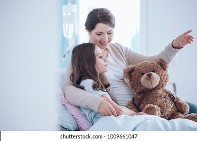 Older Sister Taking Care Of A Sick Child Lying In A Hospital Bed With Teddy Bear