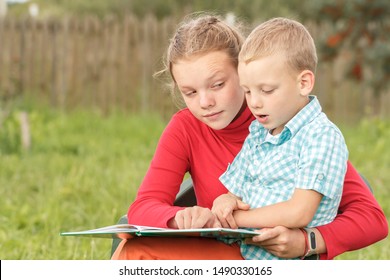 Older Sister Teaches A Little Brother To Read A Primer. Happy Children Are Studying A Book Outdoor. Mother Reading With Her Son On Nature. Toddler Sitting On His Elder Sister's Lap. Reading ABC