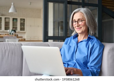 Older Smiling 60s Woman Sitting At Home On Sofa, Holding Laptop. Senior Happy Business Woman Using Pc Technologies, Working, Chatting, Spending Time In Social Media Internet, Ecommerce.