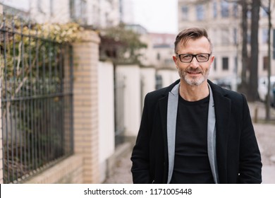 Older Stylish Man Wearing Glasses Approaching The Camera With A Quiet Smile Walking On An Urban Sidewalk