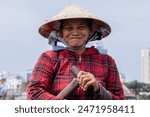 Older Vietnamese woman is looking at the camera while smiling with her mouth closed, she is on the boat with a conical hat.