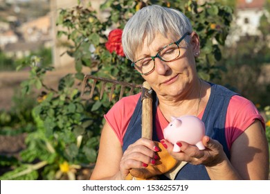 Older White Haired Woman With Glasses, Work Tool And Piggy Bank With Garden In Background