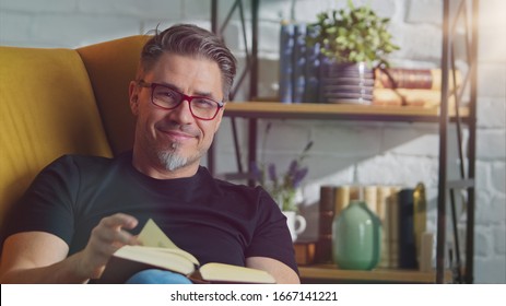 Older White Man In Glasses Reading Book At Home Sitting In Armchair In Cosy Room, Smiling.