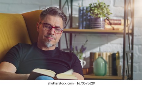 Older White Man In Glasses Reading Book At Home Sitting In Armchair In Cosy Room.