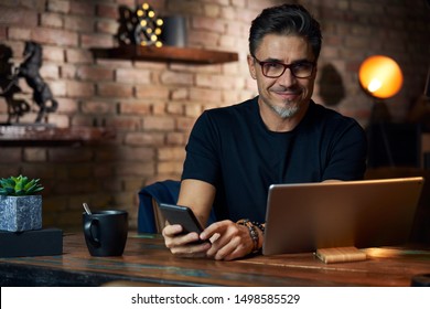 Older White Man In Glasses Sitting At Desk In Dark Room At Home, Working, Using Phone And Tablet