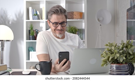 Older White Man In Glasses Sitting At Desk Using Phone. Home Office, Businessman Working From Home, Morning Coffee, Checking Social Media News Feed.