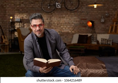 Older White Man Reading Book At Home. Wearing Casual, Gray Hair And Trendy Red Glasses.