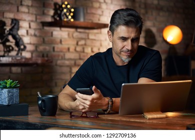 Older White Man Sitting At Desk In Dark Room At Home, Working, Using Phone And Tablet