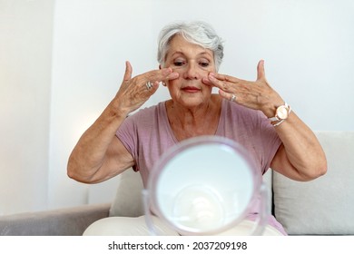 Older Woman Applying Cream To Face And Looking To Mirror At Home. Healthy Skin Care Beauty Routine Treatment Concept. Elegant Old Lady Applying Cream And Looking At Mirror In Modern Home In Sunny Day.