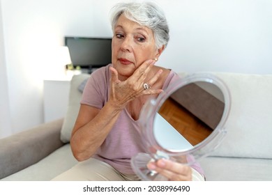 Older Woman Applying Cream To Face And Looking To Mirror At Home. Healthy Skin Care Beauty Routine Treatment Concept. Elegant Old Lady Applying Cream And Looking At Mirror In Modern Home In Sunny Day.