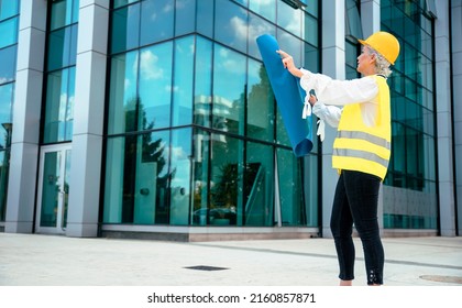 Older Woman Architect Controlling Construction Site. Elderly Female Civil Engineer With Yellow Hardhat And Reflective Vest Checking New Buildings. Senior Woman Supervisor With Blueprint. Copy Space