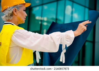 Older Woman Architect Controlling Construction Site. Elderly Female Civil Engineer With Yellow Hardhat And Reflective Vest Checking New Buildings. Senior Woman Supervisor With Blueprint. Copy Space