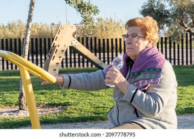 Older Woman Athlete, Drinking Water From A Bottle On The Exercise Bike In A Bio-healthy Park, At Sunset