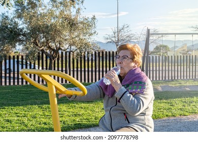 Older Woman Athlete, Drinking Water From A Bottle On The Exercise Bike In A Bio-healthy Park, At Sunset