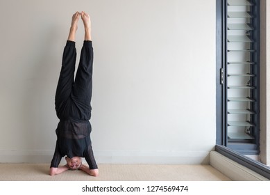 Older Woman In Black Clothing In Yoga Headstand Position Against Beige Wall (selective Focus)