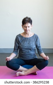 Older Woman In Black And Grey Yoga Clothing Meditating On Pink Mat