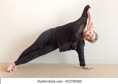 Older Woman In Black Yoga Clothing Smiling While Doing Side Yoga Posture On Floor