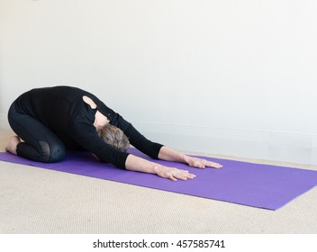 Older Woman In Black Yoga Clothing On Purple Mat In Yoga Child Posture