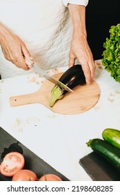 Older Woman Chef Cooking A Vegan Recipe For A Television Show. Hands Holding A Knife And Cutting An Eggplant Combined With Zucchini,tomatoes And Lettuce On A Wood Table.Balanced And Healthy Meal