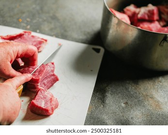 An older woman cutting a piece of meat on a cutting board. The meat is raw and has a lot of fat. Selective focus. Processing pork neck fillet for stew or barbeque. Muted colors. Food preparation - Powered by Shutterstock