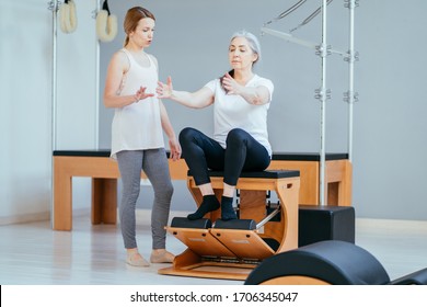 Older Woman Doing Exercise On Pilates Chair Equipment. Young Female Personal Trainer Helping Senior Woman. Workout In Rehabilitation Center, Pilates Studio. Seria Photo With Different Angles.