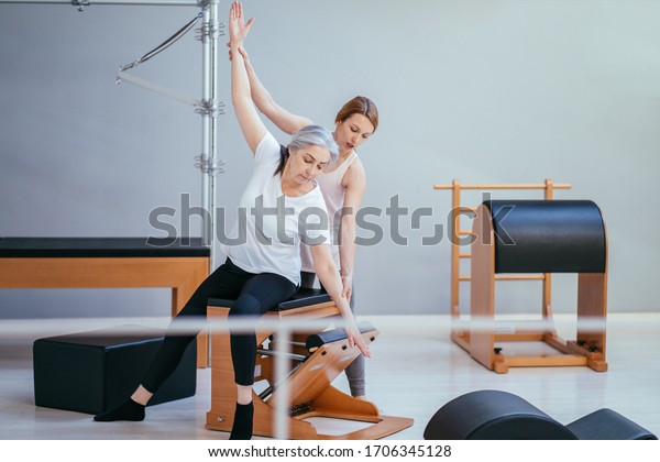 Older woman doing exercise on pilates chair\
equipment. Young female personal trainer helping senior woman.\
Workout in rehabilitation center, pilates studio. Seria photo with\
different angles.