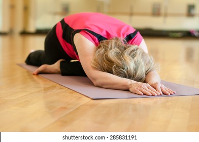 An Older Woman Doing Yoga Practices Pigeon Pose. The Blond Woman Is Wearing A Bright Pink Shirt And She Is Practicing On A Mauve Colored Yoga Mat. She Is Alone In A Studio Classroom.