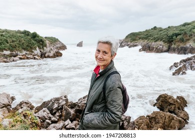 Older Woman Enjoying Her Retirement Taking A Walk On The Beach On A Cloudy Day With Strong Waves. Pensioner Looking At Camera. Active Senior People.