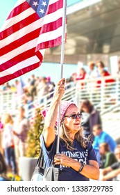 Older Woman With Flag And Womens March Tee Shirt At Political Rally With Crowd Blurred Behind Her